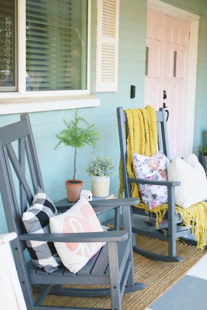 Cool grays and warm soft textures create an inviting corner to rest, crowned with a wreath that provides a spark of warm color. Piles of soft blankets make the cool summer nights feel cozy. #farmhousestyleporch #farmhouseporch #porchdecor #outdoordecor #grayrockingchairs #farmhouserockers #xbackrockingchairs