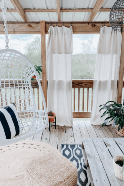 We can't decide what's the cutest part of this gorgeous boho farmhouse porch: the swing, the trendy jute circle rug or the cozy curtains. #bohofarmhouseporch #outdoorporchdecor #cozyporchideas #farmhouseporchdecor #bohostyleporch #juterugporch