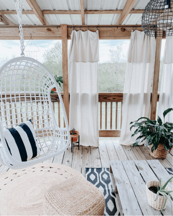 We can't decide what's the cutest part of this gorgeous boho farmhouse porch: the swing, the trendy jute circle rug or the cozy curtains. #bohofarmhouseporch #outdoorporchdecor #cozyporchideas #farmhouseporchdecor #bohostyleporch #juterugporch