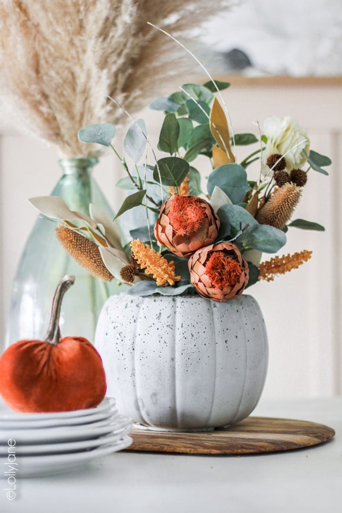 DIY Concrete Pumpkin Planter... perfect for fall and SURPRISE: the back has a jack-o-lantern face on it, perfect for Halloween! Made from a $1 treat bucket, this is a CHEAP yet CHIC project anyone can make! #diy #halloweendecor #falldecor #fallplanter #pumpkinplanter #diypumpkin #pumpkinfloralarrangement #fallfloralarrangement #fallflowers #pumpkinplanter