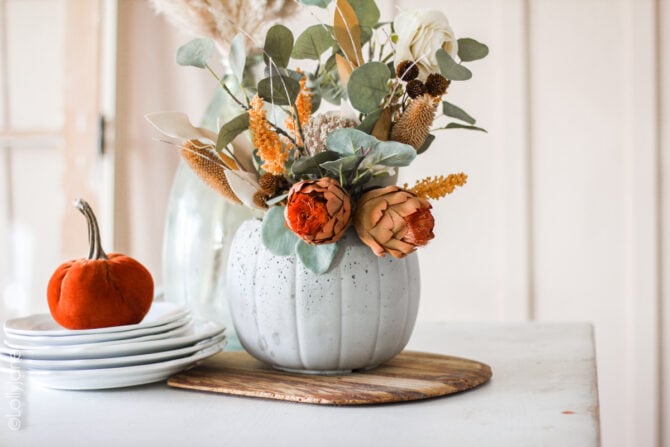 Made from a $1 treat bucket, this is a CHEAP yet CHIC project anyone can make! Darling concrete pumpkin planter. #halloweendecor #falldecor #fallplanter #pumpkinplanter #diypumpkin #pumpkinfloralarrangement #fallfloralarrangement #fallflowers #pumpkinplanter